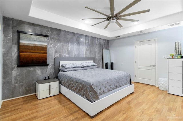 bedroom featuring a tray ceiling, ceiling fan, light hardwood / wood-style floors, and tile walls