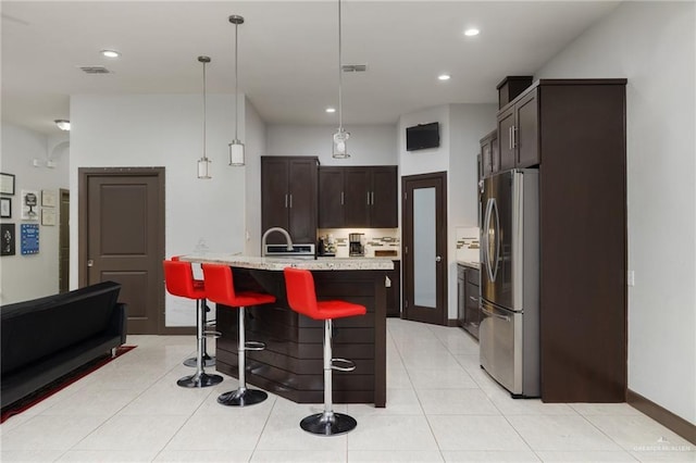 kitchen featuring pendant lighting, a center island with sink, light tile patterned floors, stainless steel refrigerator, and dark brown cabinets