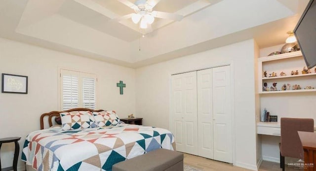 bedroom with built in desk, light hardwood / wood-style flooring, a tray ceiling, a closet, and ceiling fan