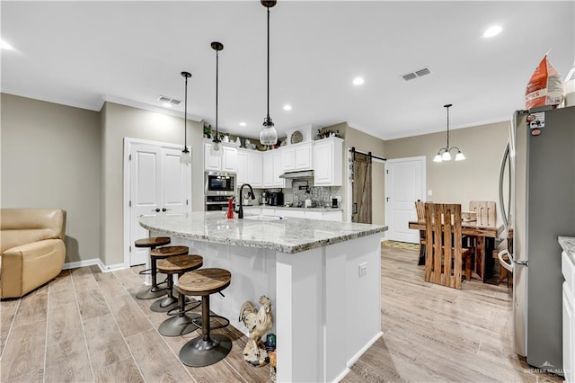 kitchen featuring light stone counters, a center island with sink, stainless steel appliances, a barn door, and white cabinets