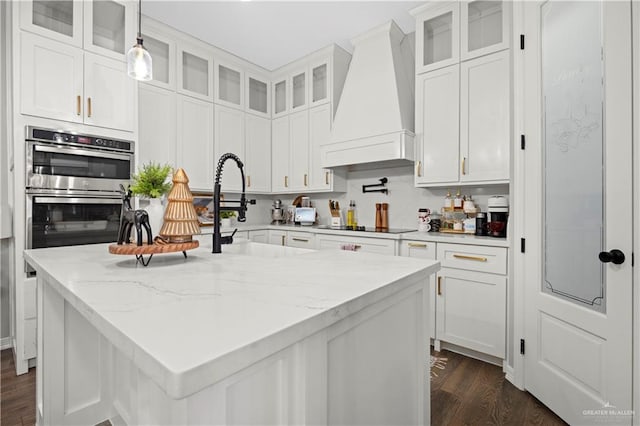 kitchen featuring white cabinetry, light stone counters, double oven, pendant lighting, and custom range hood