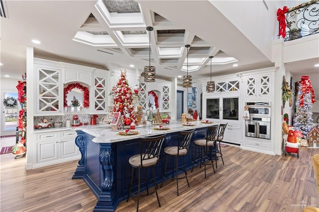 kitchen featuring white cabinetry, coffered ceiling, double oven, an island with sink, and a kitchen bar