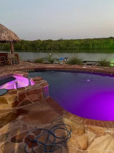 pool at dusk with a water view, an in ground hot tub, and a patio