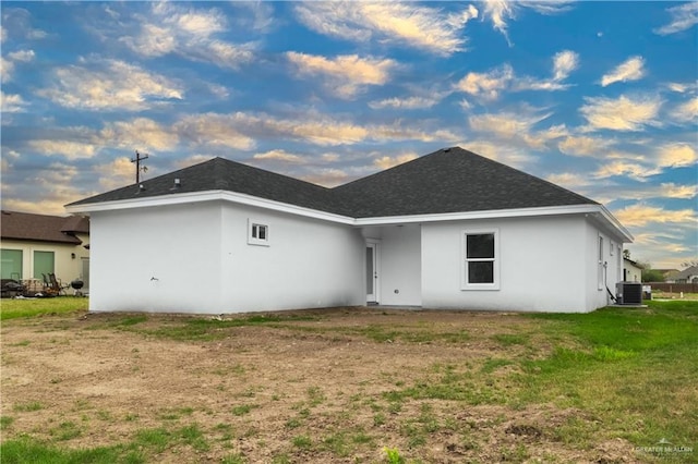 back of property at dusk featuring central air condition unit, a lawn, a shingled roof, and stucco siding