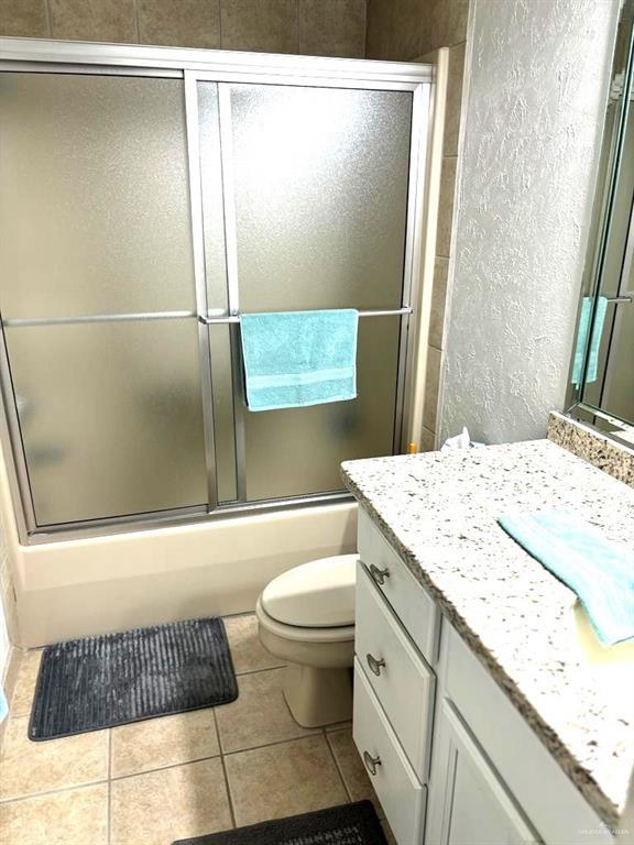 bathroom featuring bath / shower combo with glass door, toilet, a textured wall, tile patterned floors, and vanity