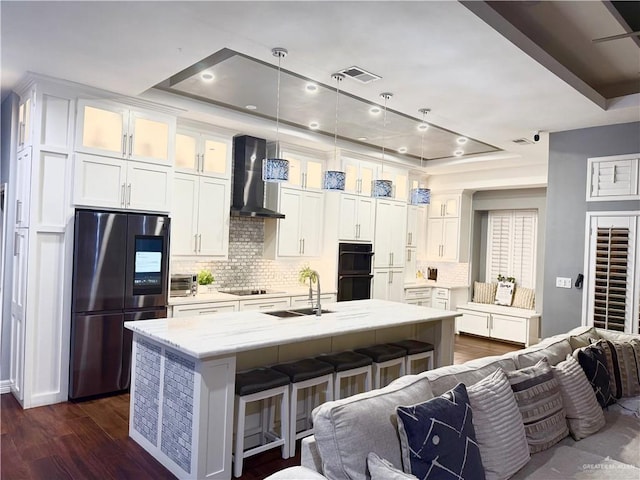 kitchen featuring an island with sink, a tray ceiling, black appliances, and wall chimney range hood