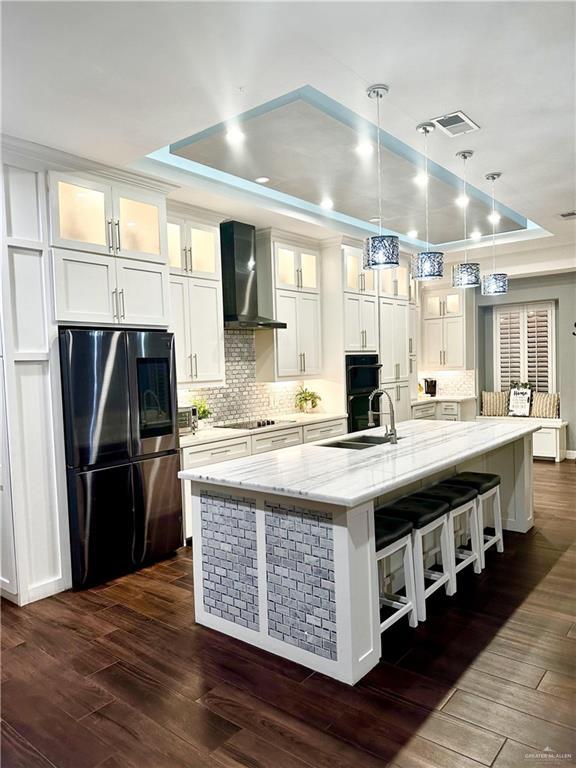 kitchen featuring wall chimney exhaust hood, stainless steel fridge, a raised ceiling, and an island with sink