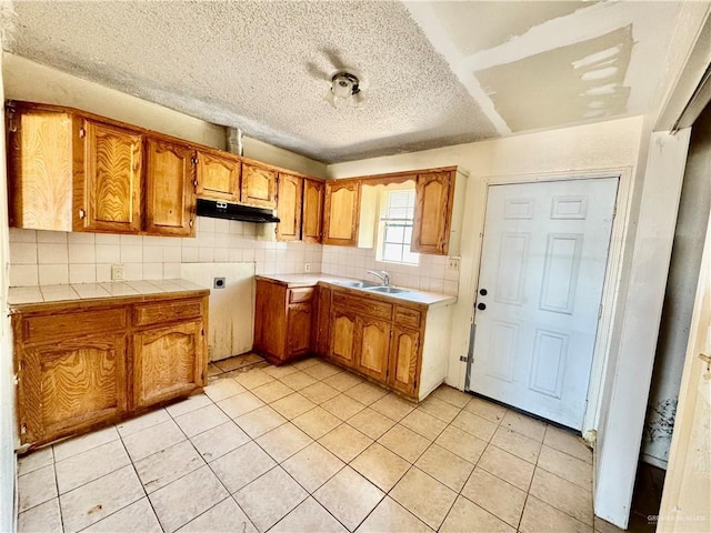 kitchen featuring under cabinet range hood, backsplash, brown cabinetry, and a textured ceiling