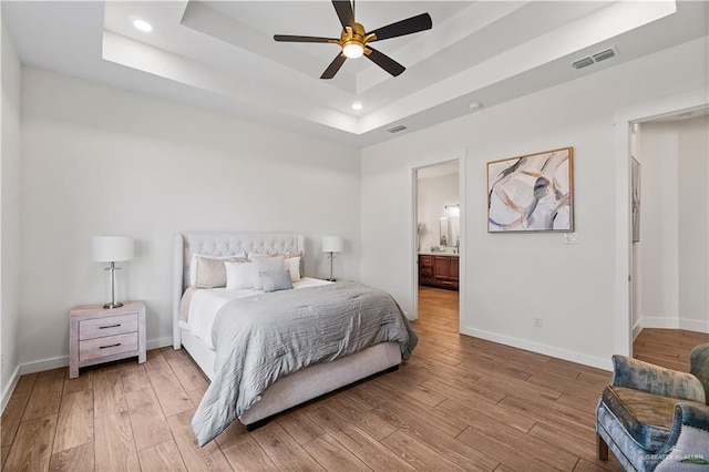 bedroom with ensuite bathroom, a raised ceiling, ceiling fan, and light wood-type flooring