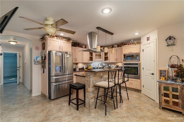 kitchen with arched walkways, stainless steel appliances, a kitchen breakfast bar, wall chimney exhaust hood, and tasteful backsplash