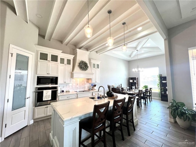 kitchen featuring an island with sink, stainless steel oven, built in microwave, white cabinets, and beamed ceiling