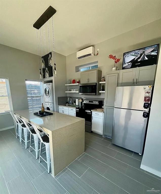 kitchen featuring pendant lighting, open shelves, stainless steel appliances, stacked washer and dryer, and an island with sink