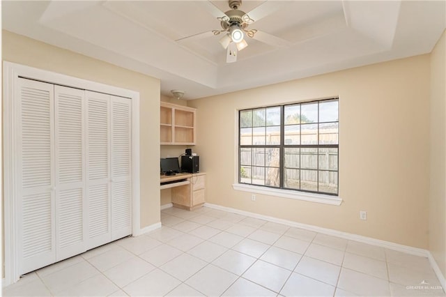 unfurnished office with light tile patterned flooring, built in desk, ceiling fan, and a tray ceiling