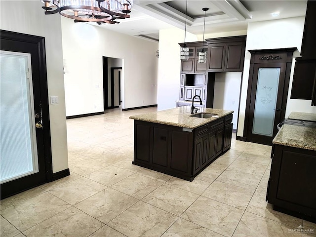 kitchen with dark brown cabinetry, sink, light stone counters, an island with sink, and pendant lighting