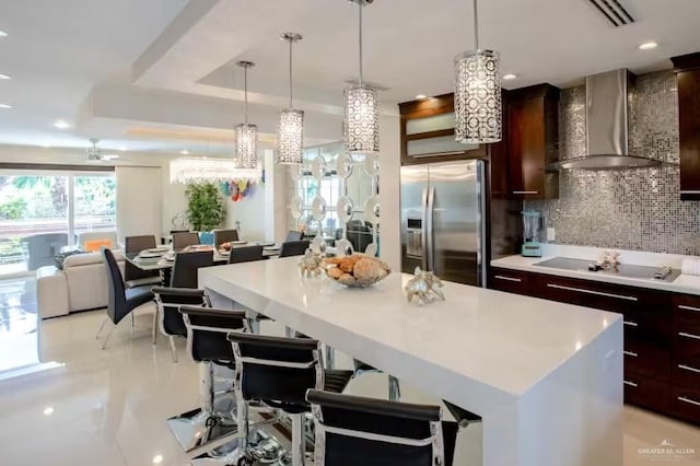 kitchen featuring a raised ceiling, light countertops, wall chimney range hood, stainless steel fridge, and black electric cooktop