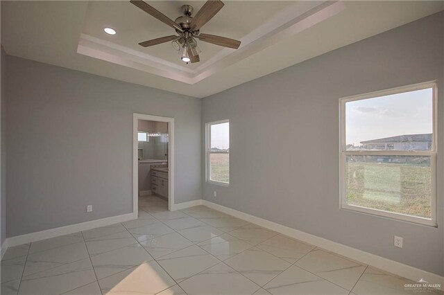 unfurnished bedroom featuring ensuite bath, ceiling fan, a tray ceiling, and multiple windows