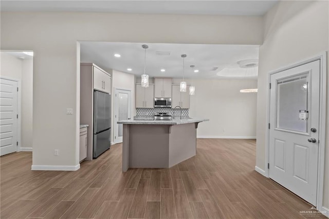 kitchen with appliances with stainless steel finishes, light wood-type flooring, backsplash, a kitchen island with sink, and pendant lighting