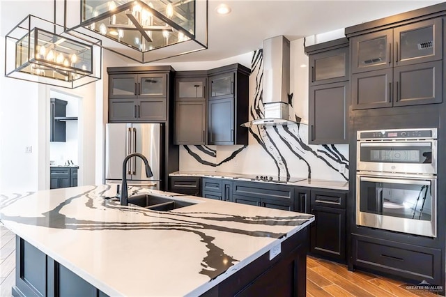 kitchen featuring stainless steel appliances, a sink, light countertops, wall chimney exhaust hood, and glass insert cabinets