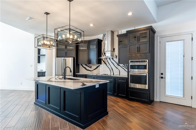 kitchen featuring visible vents, an island with sink, appliances with stainless steel finishes, wall chimney range hood, and pendant lighting