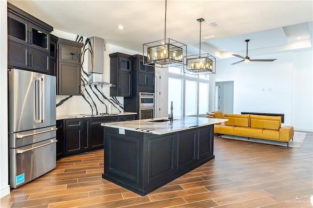 kitchen featuring light stone counters, pendant lighting, stainless steel appliances, a kitchen island with sink, and dark cabinets