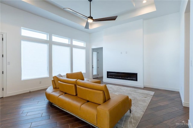 living room with a tray ceiling, dark wood-style flooring, a glass covered fireplace, and a ceiling fan