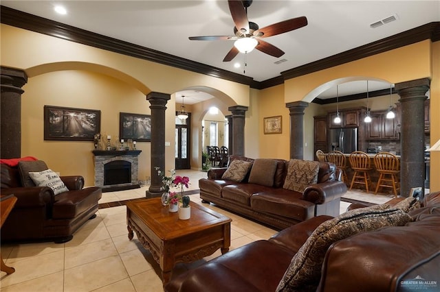 tiled living room with decorative columns, a stone fireplace, ceiling fan, and crown molding