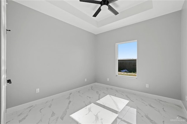 empty room with a tray ceiling, marble finish floor, baseboards, and a ceiling fan