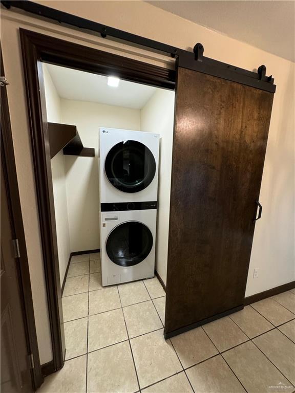 laundry room featuring a barn door, stacked washer / dryer, light tile patterned floors, and baseboards