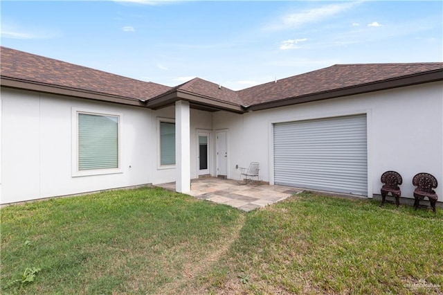 rear view of property featuring a shingled roof, stucco siding, a yard, an attached garage, and a patio