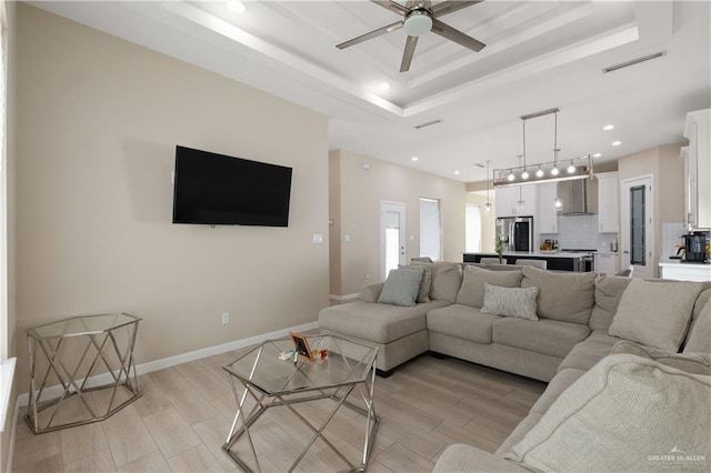 living area featuring baseboards, light wood-style flooring, recessed lighting, ceiling fan, and a raised ceiling