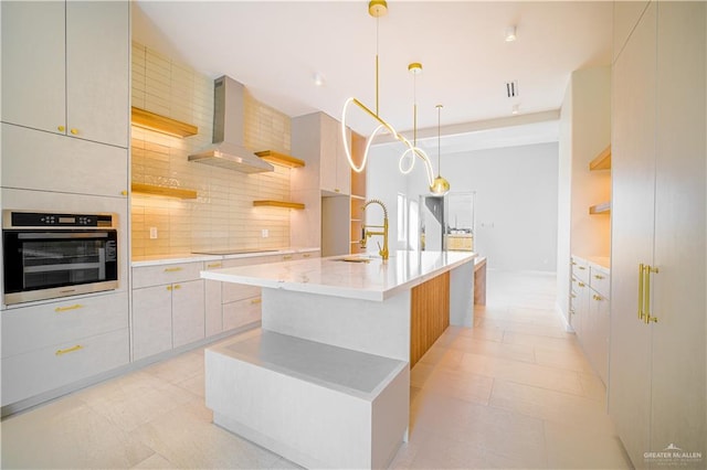 kitchen featuring sink, wall chimney range hood, oven, pendant lighting, and a center island with sink