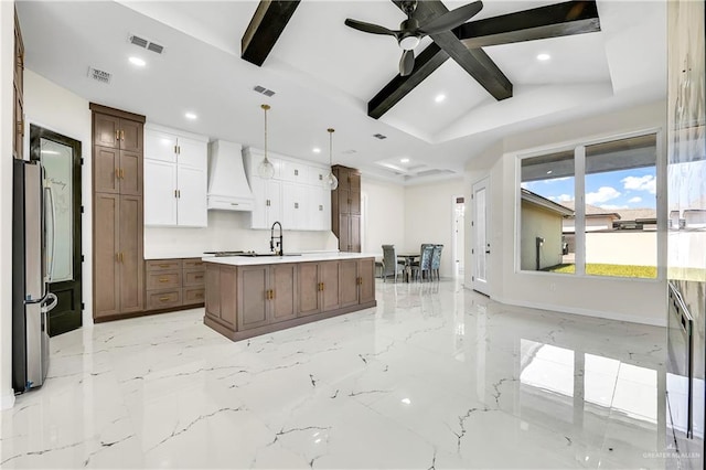 kitchen featuring a kitchen island with sink, hanging light fixtures, custom range hood, white cabinetry, and stainless steel refrigerator