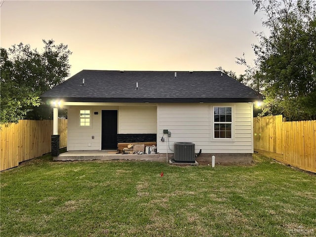 back of property at dusk with a yard, a patio area, a fenced backyard, and a shingled roof