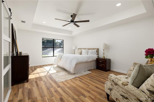 bedroom with a tray ceiling, light hardwood / wood-style flooring, ceiling fan, and a barn door