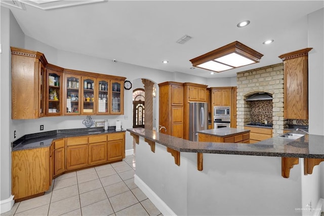 kitchen with a breakfast bar area, stainless steel appliances, dark countertops, glass insert cabinets, and a sink