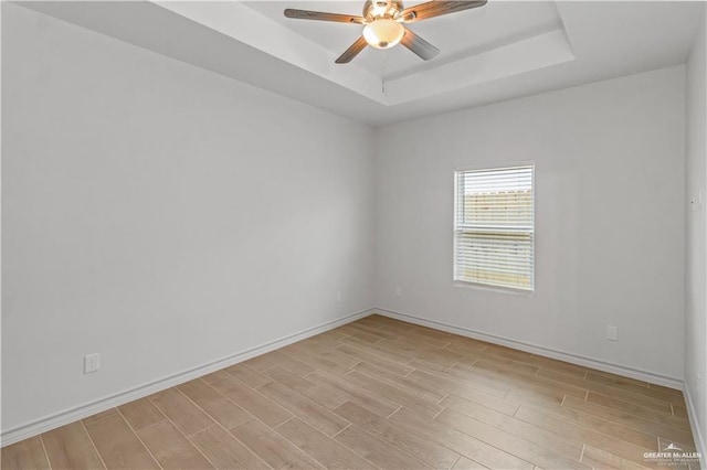 empty room featuring a tray ceiling, light wood-style flooring, baseboards, and ceiling fan