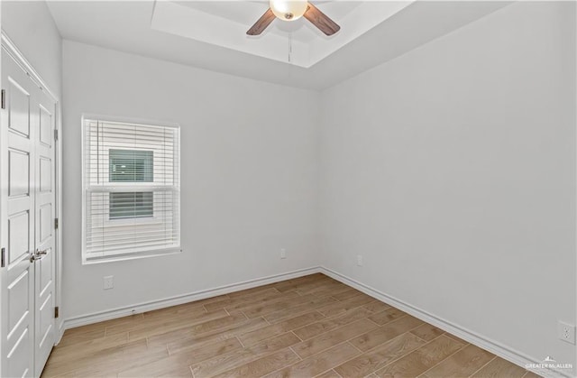 empty room featuring a raised ceiling, baseboards, light wood-type flooring, and ceiling fan