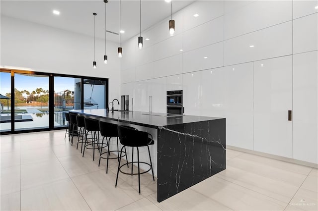 kitchen featuring a breakfast bar, hanging light fixtures, a large island with sink, dark stone counters, and white cabinets