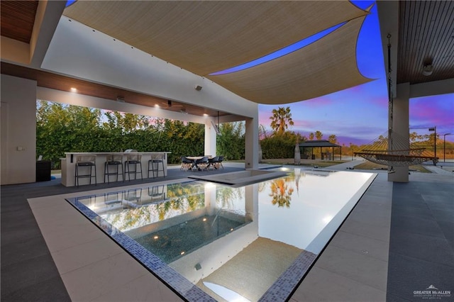 pool at dusk with a gazebo, ceiling fan, an outdoor bar, and a patio