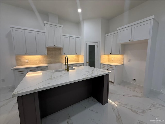 kitchen with a sink, a center island with sink, light stone countertops, backsplash, and baseboards