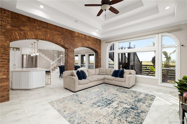 living room with a tray ceiling, arched walkways, marble finish floor, and a ceiling fan