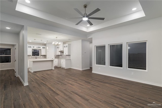 unfurnished living room with a tray ceiling, dark wood-style flooring, baseboards, and ceiling fan with notable chandelier