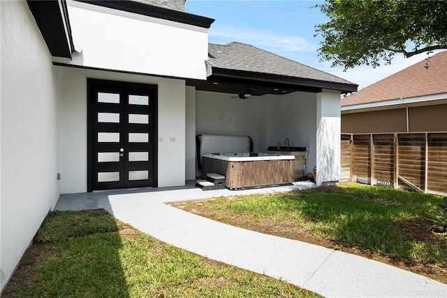 doorway to property featuring a ceiling fan, stucco siding, fence, a hot tub, and a shingled roof