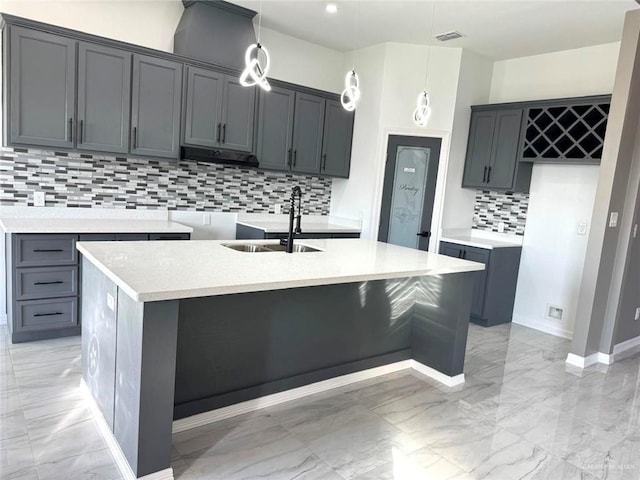 kitchen with tasteful backsplash, sink, hanging light fixtures, gray cabinets, and a center island with sink