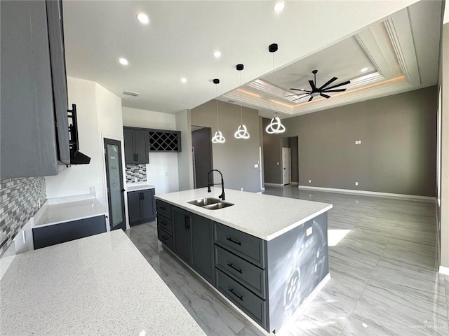 kitchen featuring backsplash, sink, a tray ceiling, hanging light fixtures, and a kitchen island with sink