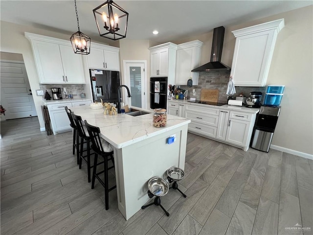 kitchen featuring wall chimney exhaust hood, pendant lighting, a center island with sink, black appliances, and white cabinetry