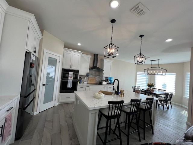 kitchen with a kitchen island with sink, black appliances, pendant lighting, wall chimney exhaust hood, and white cabinets