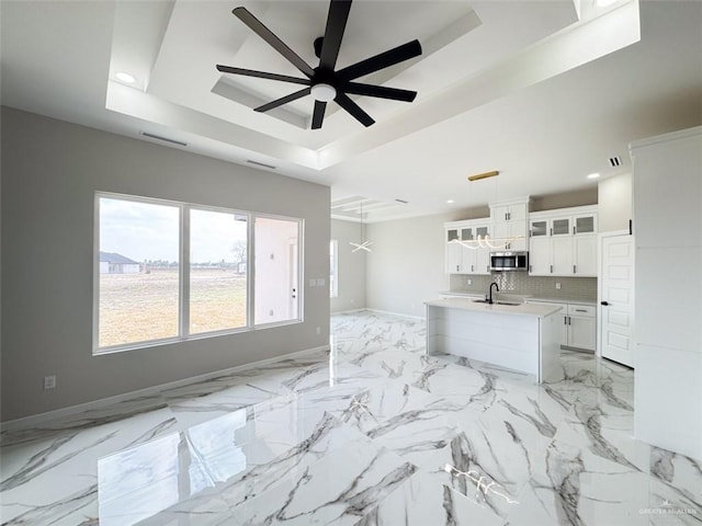 kitchen featuring pendant lighting, an island with sink, white cabinets, backsplash, and a raised ceiling