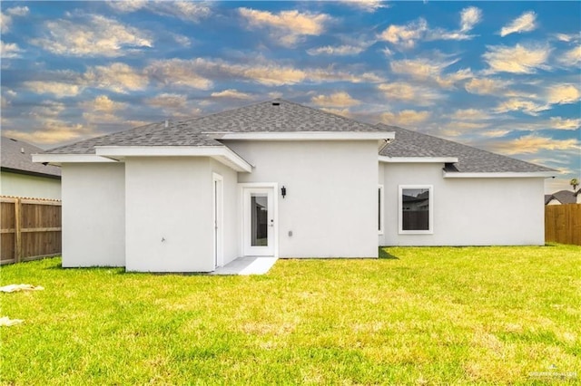 rear view of property featuring roof with shingles, fence, a lawn, and stucco siding