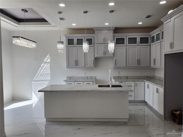 kitchen featuring a kitchen island with sink, sink, hanging light fixtures, gray cabinets, and light stone countertops